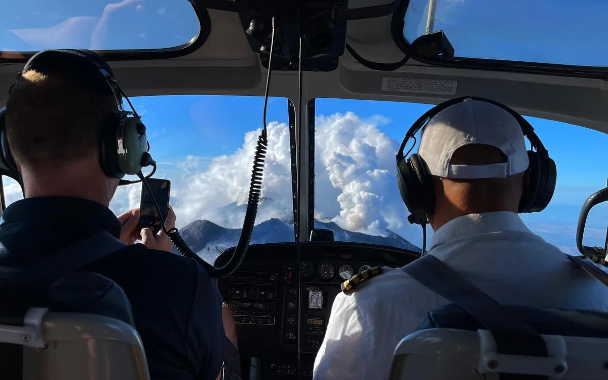 A helicopter pilot in the left-hand seat with both hands on the controls and the instrument panel lit in front of him